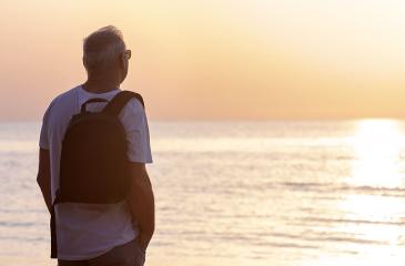 An adult man with a backpack on his shoulder admires the sunset on the sea standing with his back to the viewer